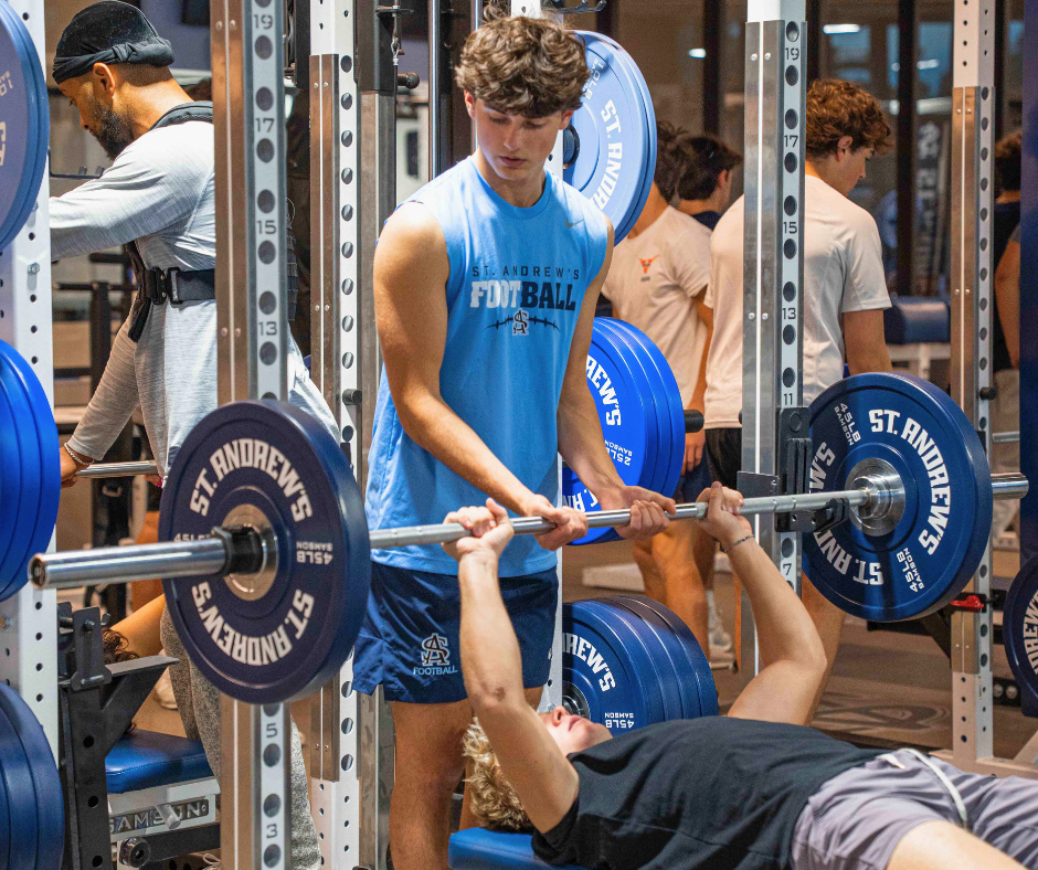 St. Andrew's football player helps another player bench press a large weight.