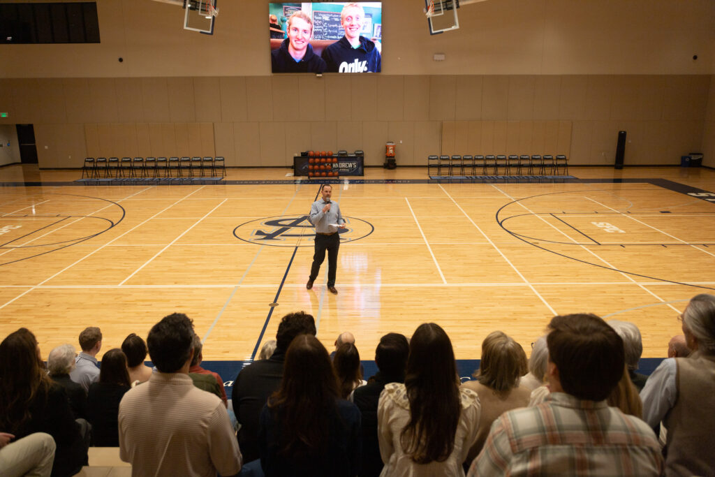 Coach speaks into a microphone while facing a crowd. A photo of Johnson and his brother is on the screen behind him.