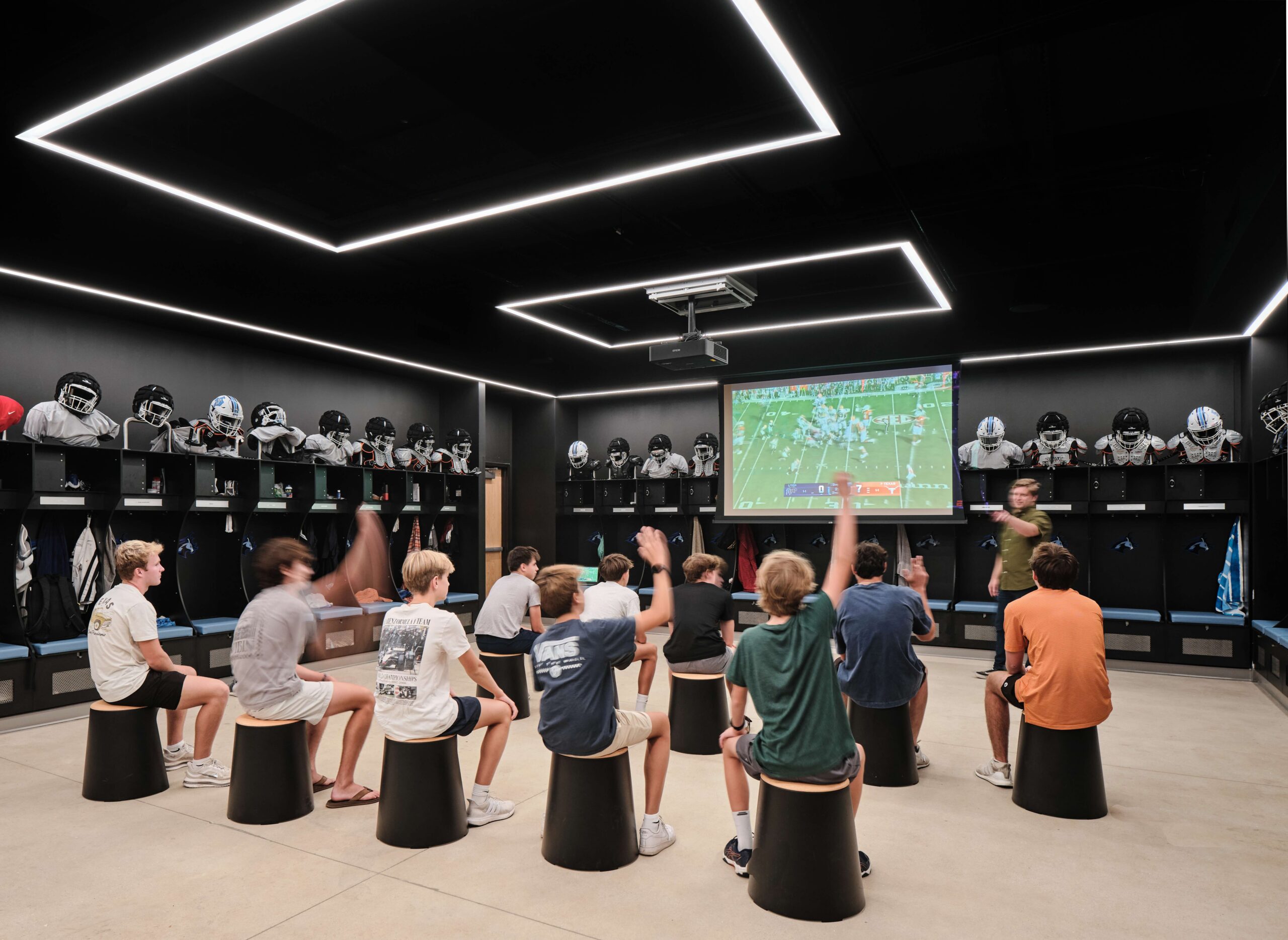 Students watch footage on a large screen in a locker room.