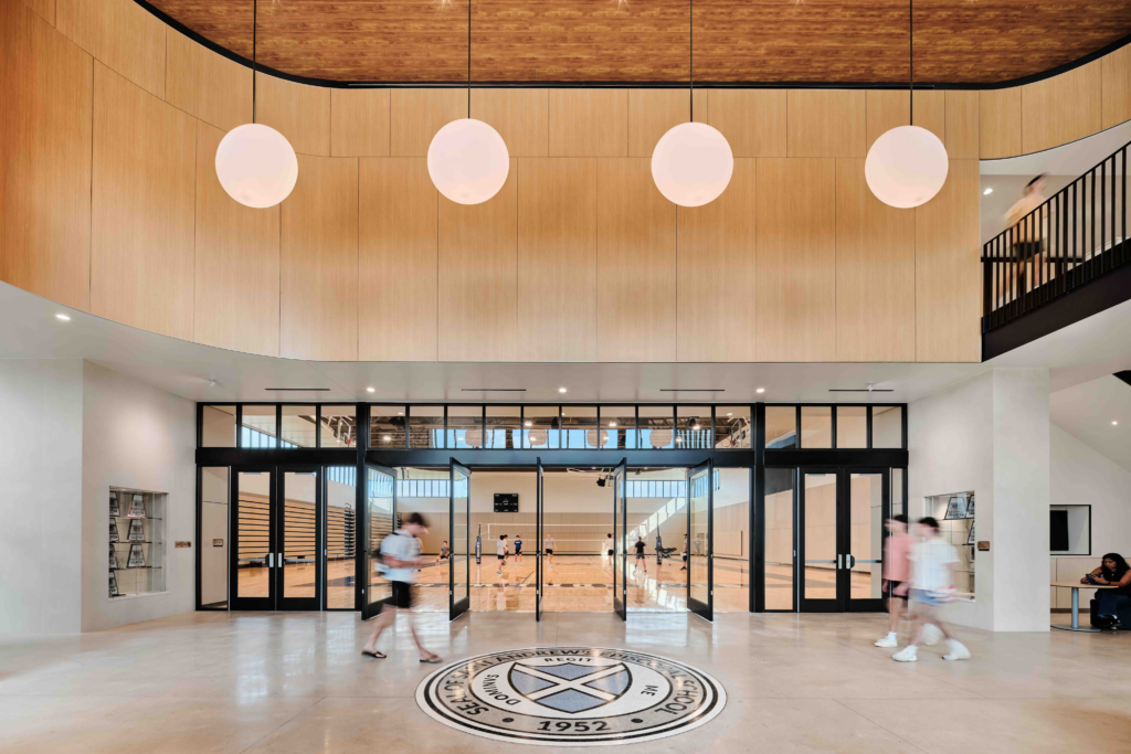 A spacious, modern school lobby featuring a large circular floor emblem for St. Andrew's Episcopal School. The room has tall wood-paneled walls, spherical pendant lights, and large glass doors that provide a view into a bright gymnasium.