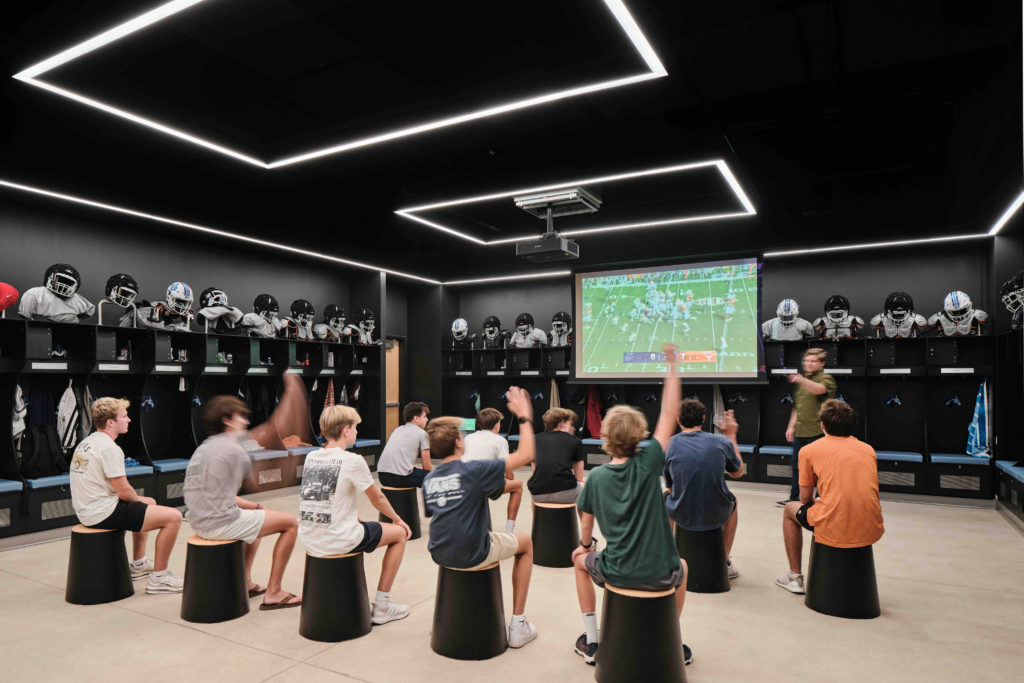 A dark, modern locker room with black walls and built-in shelving displaying football helmets and jerseys. A group of students is seated on black stools, watching a football game projected onto a large screen under rectangular LED ceiling lights.