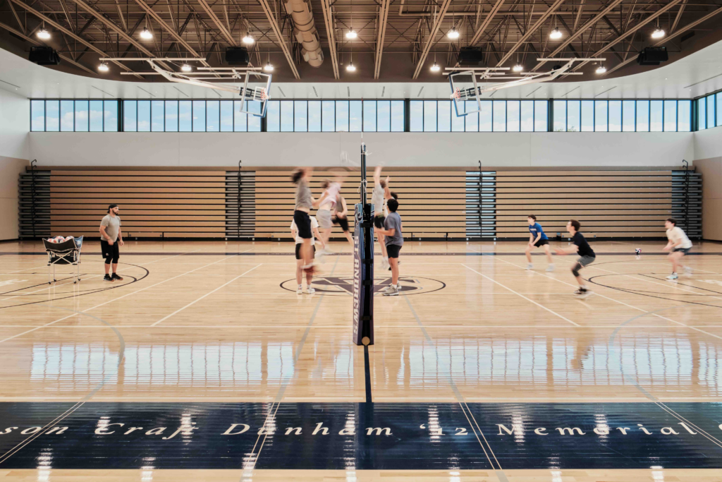 An indoor volleyball match in a large gymnasium with polished wood floors. Players are captured in mid-action near a net labeled "ST. ANDREW'S," with tiered bleachers and a row of high windows in the background.