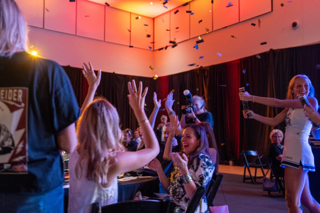 People sitting at a table cheer as SAS cheerleaders shoot confetti from confetti cannons.