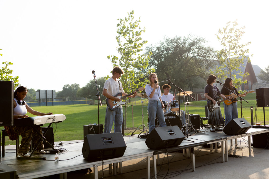Students play their rock band instruments while standing on a stage outside.